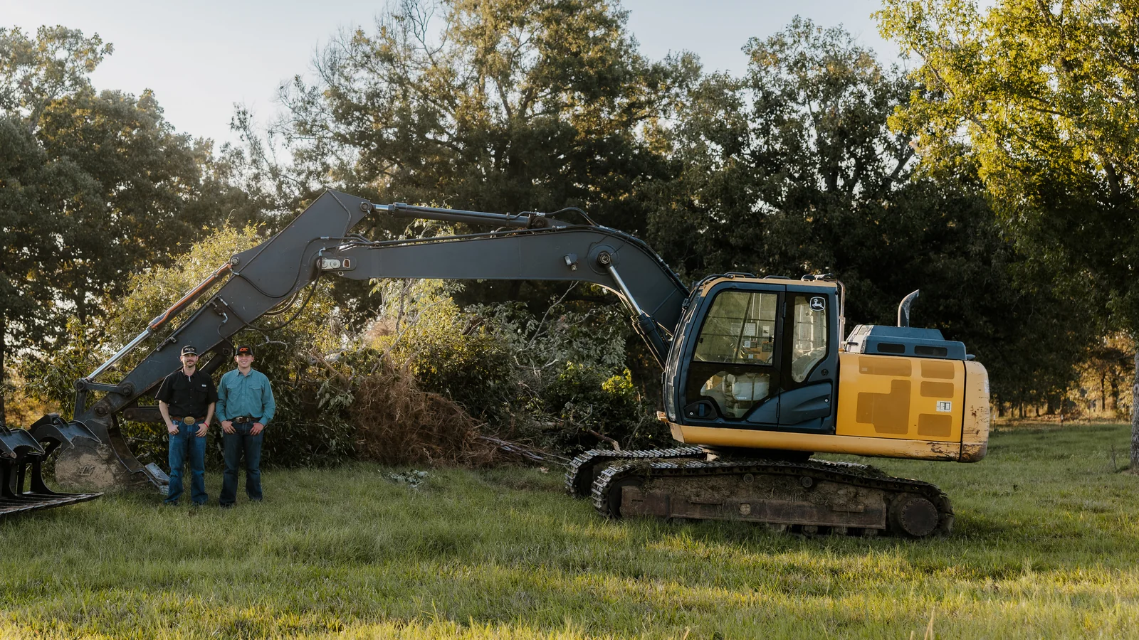 Luke and Grady beside their excavator and a cleared brush pile at sunset